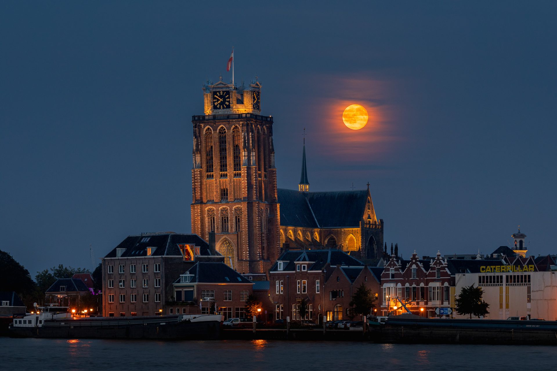 A full orange moon rising next to the illuminated tower of the Grote Kerk in Dordrecht, seen across the river at dusk
